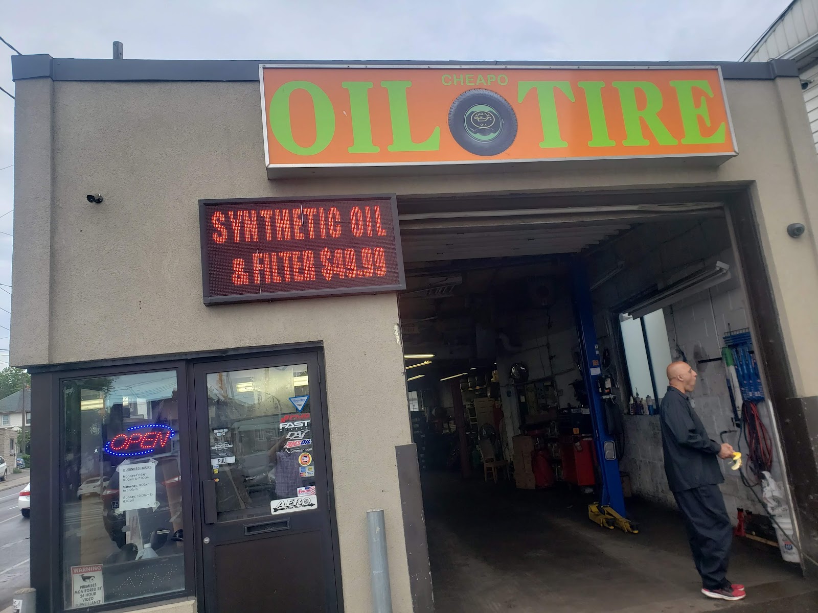 The Cheapo Oil and Tire storefront on Rogers Rd: bright orange marquee with the shop name, an LED sign reading SYNTHETIC OIL & FILTER $49.99, and an open garage bay.